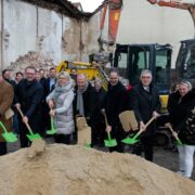 Feierlicher Spatenstich für das Haus Lichtermeer: Eine Gruppe von Beteiligten steht mit leuchtend grünen Schaufeln an einem Sandhaufen auf dem künftigen Baugelände, im Hintergrund sind weitere Gäste und ein Bagger zu sehen.