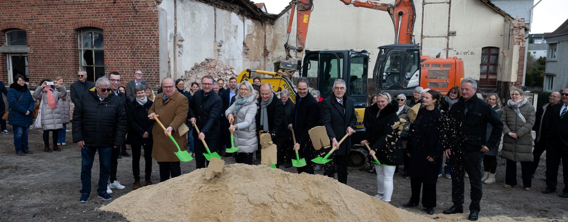Feierlicher Spatenstich für das Haus Lichtermeer: Eine Gruppe von Beteiligten steht mit leuchtend grünen Schaufeln an einem Sandhaufen auf dem künftigen Baugelände, im Hintergrund sind weitere Gäste und ein Bagger zu sehen.